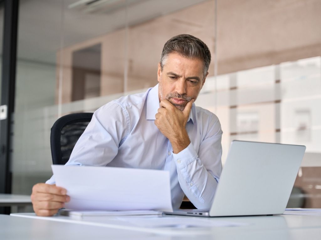 Empresario sentado frente a un computador en una oficina, observa una hoja de papel con cara pensativa