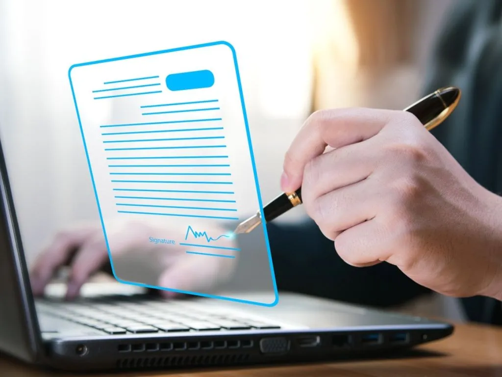 Una mano con un lápiz simula hacer una firma electrónica sobre una proyección virtual de un documento