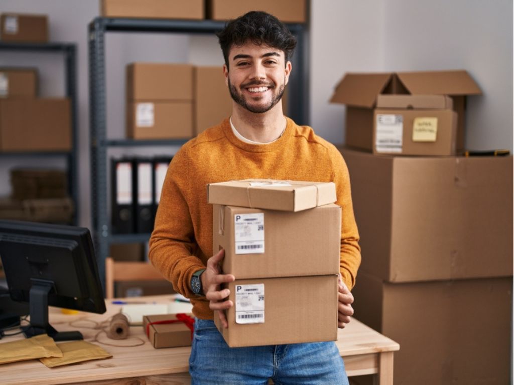 Una persona sonríe con cajas en las manos mientras se apoya en un escritorio que detrás presenta estantes con más cajas de sus emprendimientos.