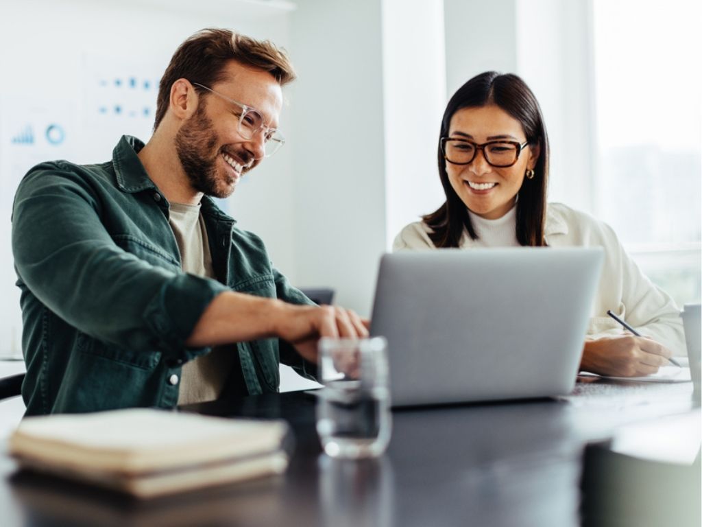Un hombre y una mujer están sentados frente a una computadora mientras sonríen al leer los fondos para emprendedores a los que pueden postular.
