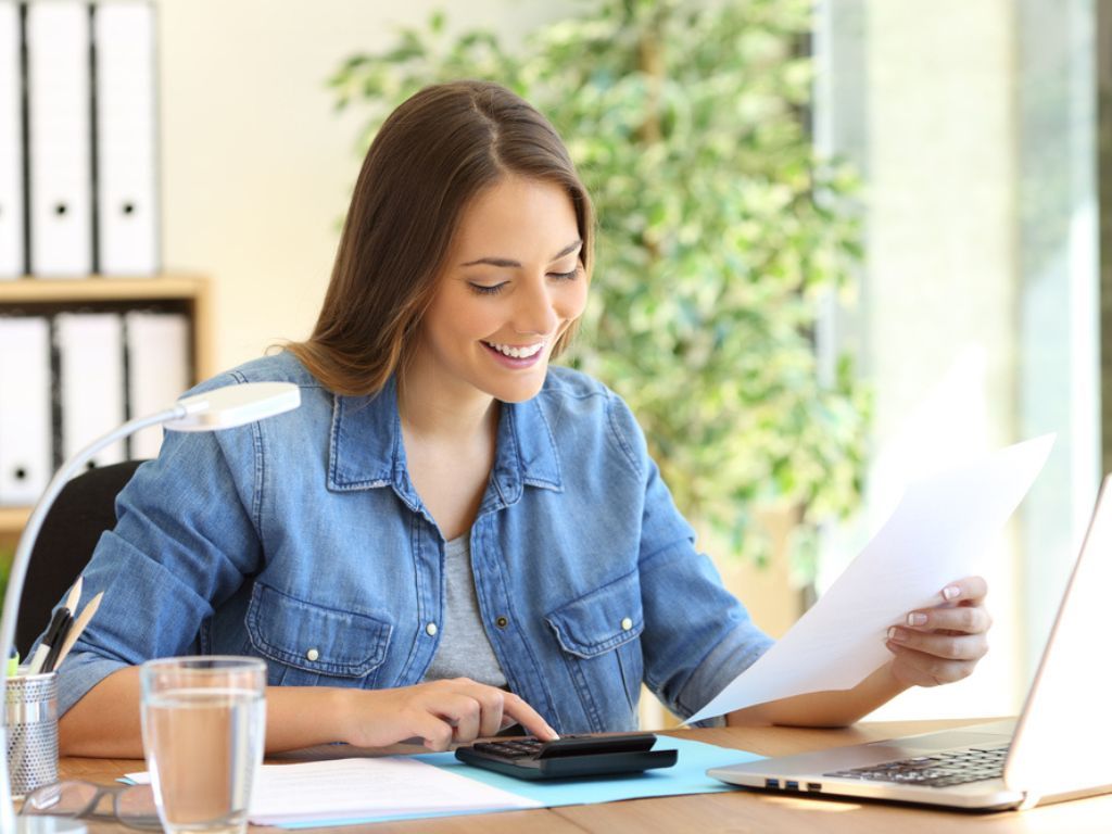 Una mujer sonríe porque realiza cómodamente la operación renta 2024 desde su computadora, además sostiene un documento en una mano y utiliza una calculadora con la otra.