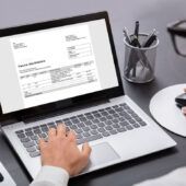 Se muestra un escritorio con una persona que calcula a mano una factura en papel, en frente hay un computador con la misma factura, ya que está explorando qué es una factura electrónica y sus beneficios.