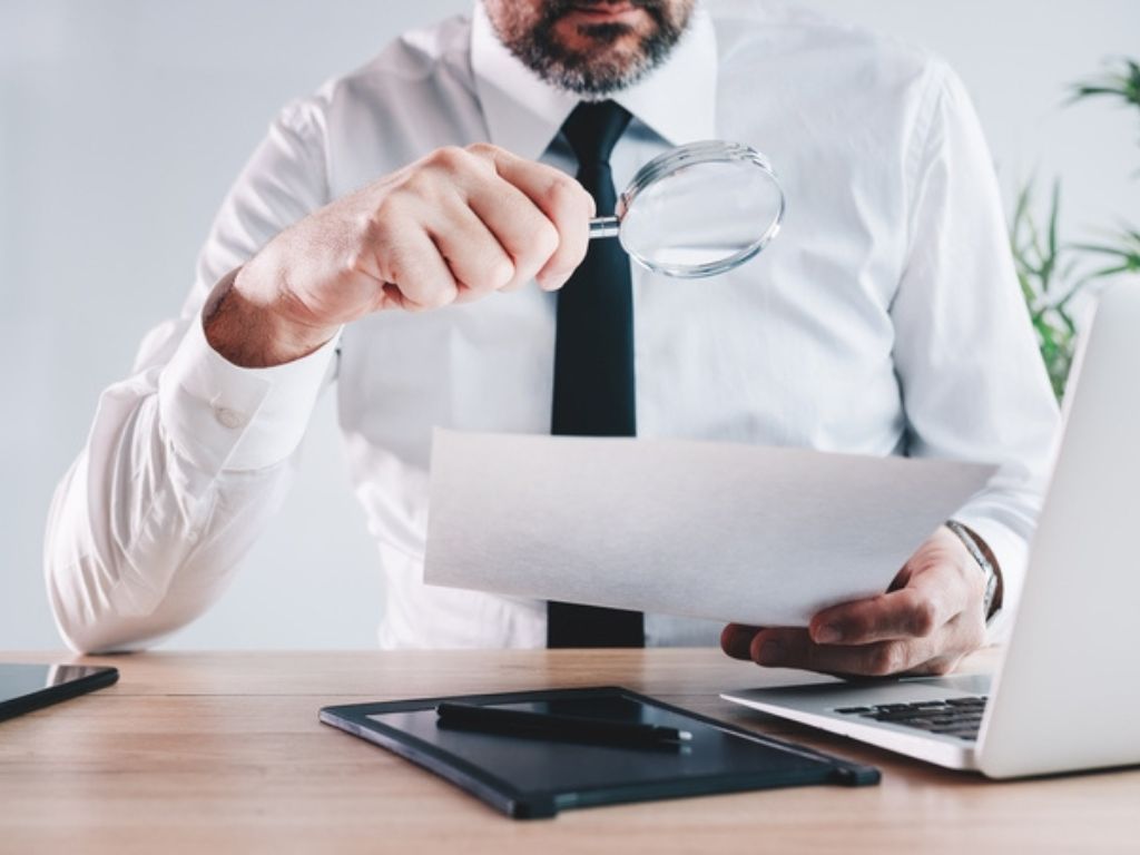 Un hombre sentado en un escritorio sostiene una lupa con la que observa un documento, representando la búsqueda de servicios de contabilidad.